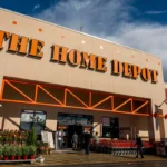Home Depot store exterior with orange signage, American flags, shoppers and carts under a cloudy sky.