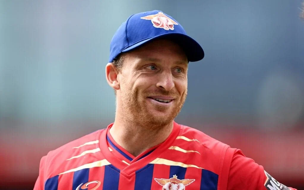 Jos Buttler smiles in a Red Bull cricket jersey and blue cap during a T20 cricket match, proudly.