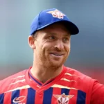 Jos Buttler smiles in a Red Bull cricket jersey and blue cap during a T20 cricket match, proudly.