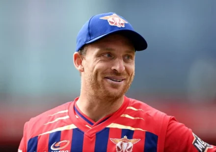 Jos Buttler smiles in a Red Bull cricket jersey and blue cap during a T20 cricket match, proudly.