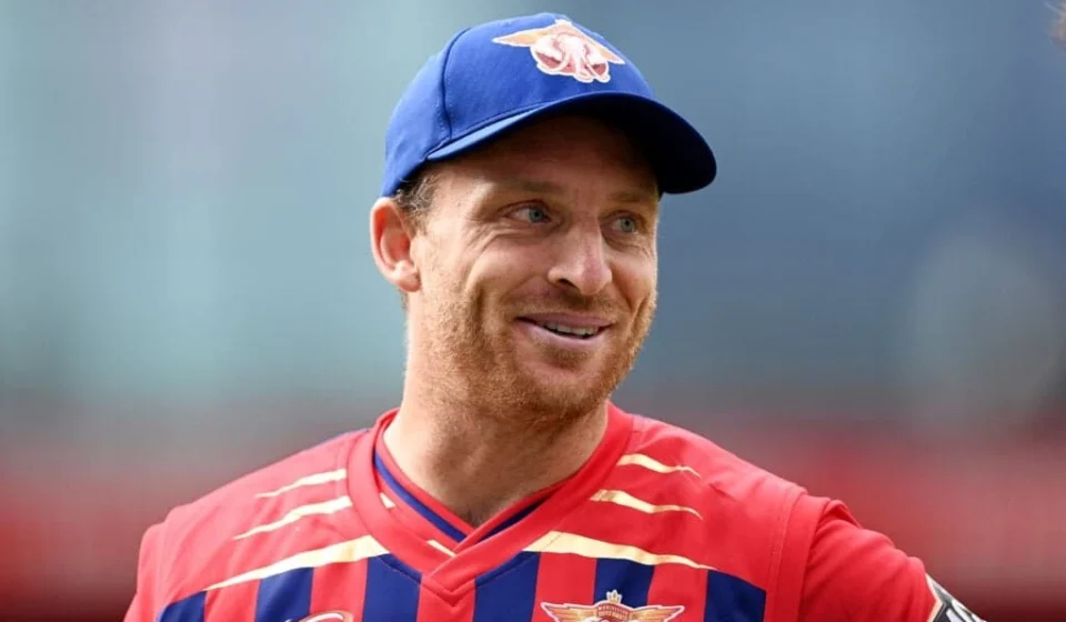 Jos Buttler smiles in a Red Bull cricket jersey and blue cap during a T20 cricket match, proudly.