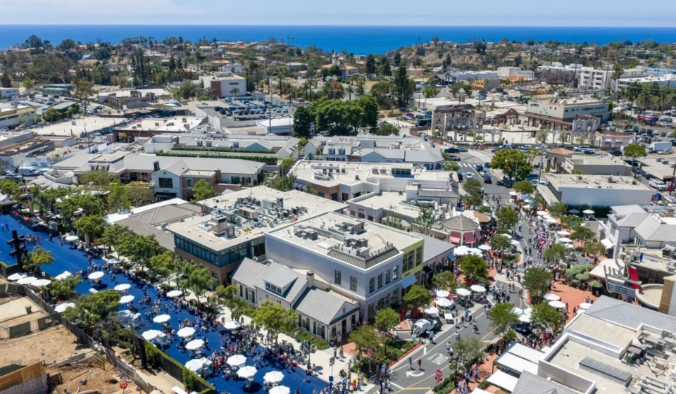 Palisades Village Hub Reopens Following Fire Restoration Aerial view of Palisades Village with shops, streets, crowds, and the Pacific Ocean in the distance.