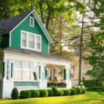 Green and white cottage home with wraparound porch, shaded by trees