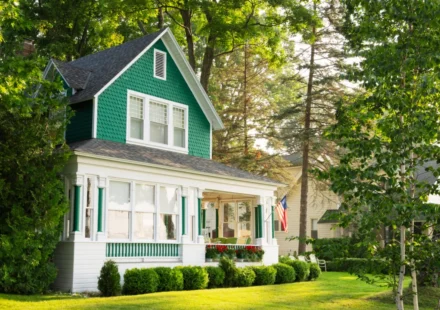 Green and white cottage home with wraparound porch, shaded by trees