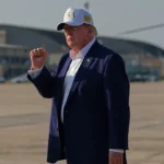 President Trump raises his fist while standing on an airport tarmac wearing a dark suit and USA cap.