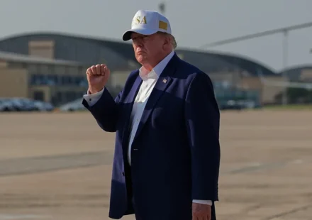 President Trump raises his fist while standing on an airport tarmac wearing a dark suit and USA cap.