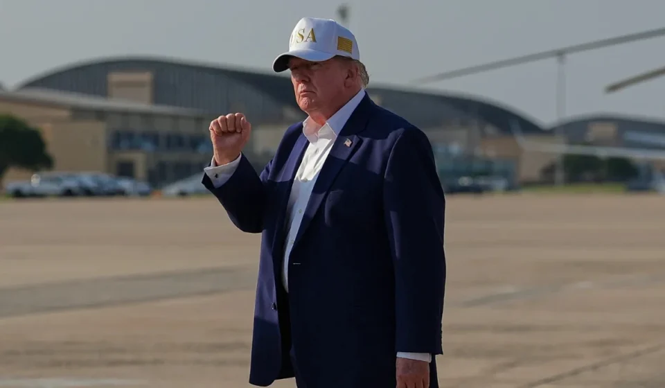 President Trump Demands Conflict Compensation from Iran President Trump raises his fist while standing on an airport tarmac wearing a dark suit and USA cap.