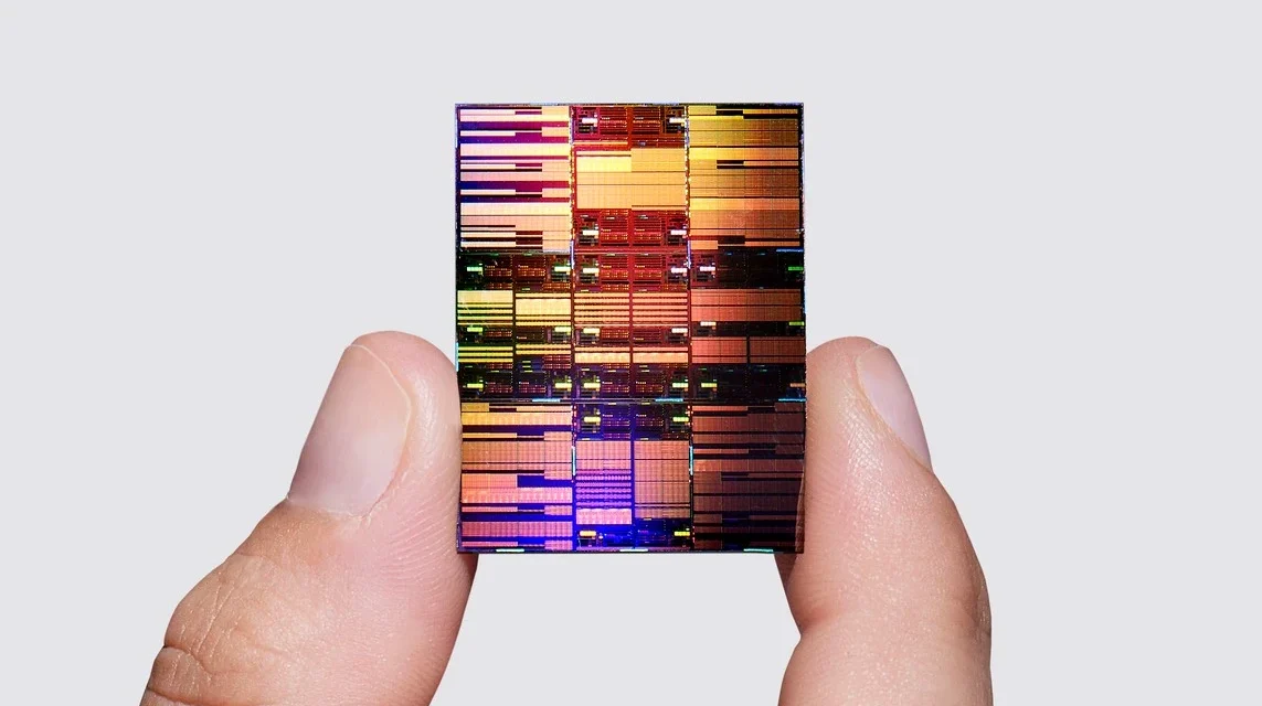 A semiconductor chip held between fingers displays a detailed multicolored circuit pattern up close.