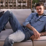Model lounges on rooftop sofa wearing light blue denim shirt and jeans for Fall fashion campaign fit