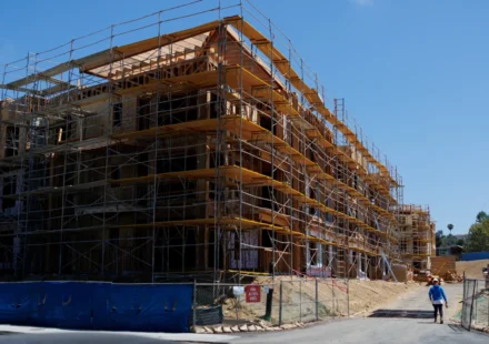 Construction workers walk past a multi story residential building under construction with scaffolding.