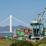 Cargo containers and cranes at a busy US port beneath a suspension bridge reflecting global trade.