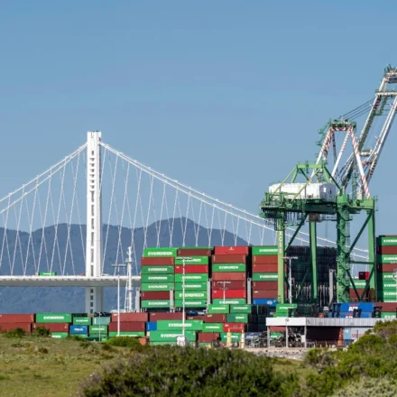 Cargo containers and cranes at a busy US port beneath a suspension bridge reflecting global trade.