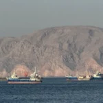 Cargo ships sail through the Strait of Hormuz with rocky coastline during US Iran maritime talks underway.