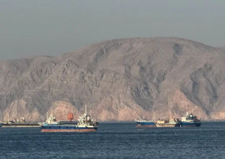 Cargo ships sail through the Strait of Hormuz with rocky coastline during US Iran maritime talks underway.